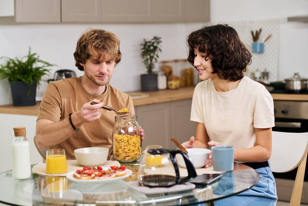 Młody człowiek biorąc płatki kukurydziane ze słoika przygotowując śniadanie przy stole kuchennym przed żoną z misą patrząc na niego - Zdjęcie, obraz
