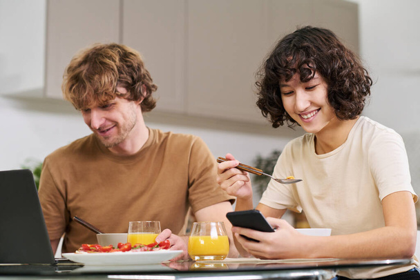 Jovem feliz com smartphone comendo flocos de milho com leite enquanto sentada à mesa da cozinha ao lado de seu marido em rede na frente do laptop - Foto, Imagem