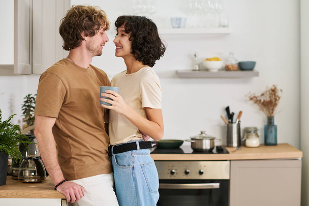 Счастливые молодые возлюбленные в повседневной одежде, стоящие в объятиях перед камерой на кухне, в то время как веселая женщина пьет кофе - Фото, изображение