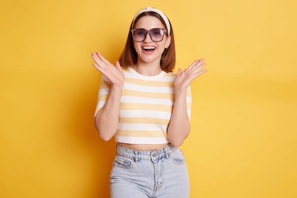 Pleasantly surprised woman wearing striped T-shirt hair band and sunglasses standing isolated over yellow background raised her arms with happy positive expression. - Foto, imagen