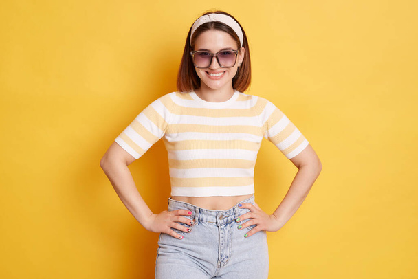 Confident cheerful woman wearing striped T-shirt hair band and sunglasses standing isolated over yellow background posing with hands on hips being in good mood. - Zdjęcie, obraz