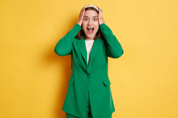 Shocked surprised woman wearing green jacket standing isolated over yellow background keeps hands on her head screaming with amazed expression. - Valokuva, kuva