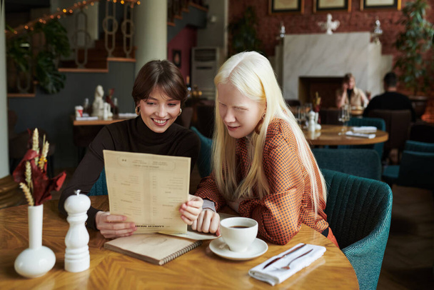 Albino κορίτσι και χαρούμενος φίλος της κοιτάζοντας μέσα από το μενού και την επιλογή των τροφίμων, ενώ κάθεται δίπλα στο τραπέζι σε ζεστό καφέ κατά τη διάρκεια της συνάντησής τους στον ελεύθερο χρόνο - Φωτογραφία, εικόνα