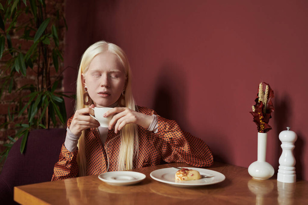 Young restful albino woman in smart casual attire enjoying craft coffee with tasty dessert while spending time in cozy cafe with maroon walls - Photo, image