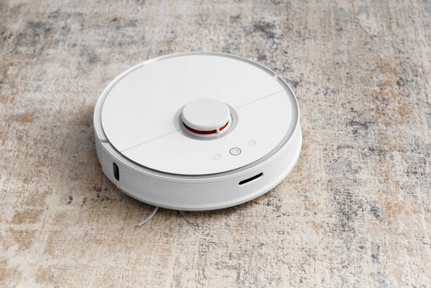 White robot vacuum cleaner on the carpet in the living room near the sofa. Cleaning the apartment with a modern smart vacuum cleaner. - Photo, Image