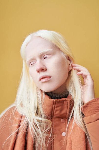 Joven mujer albina serena con el pelo largo y rubio de pie delante de la cámara en aislamiento contra la pared amarilla del edificio moderno - Foto, Imagen