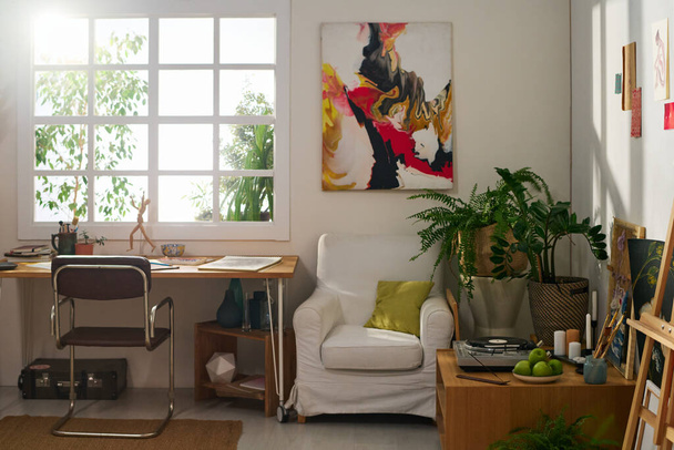 Рабочее место современного художника у окна просторной домашней студии со столом, стулом, креслом, абстрактной живописью на стене и зелеными растениями - Фото, изображение