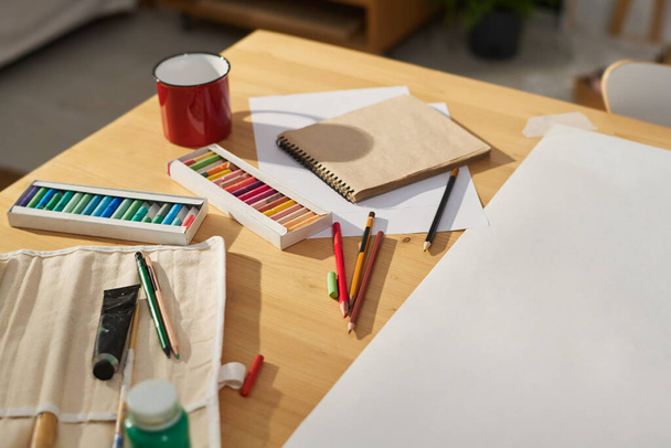 Parte de la mesa con taza pequeña, lápices de colores múltiples, pinturas, cuaderno y hoja de papel en blanco necesarios para dibujar bocetos creativos - Foto, Imagen