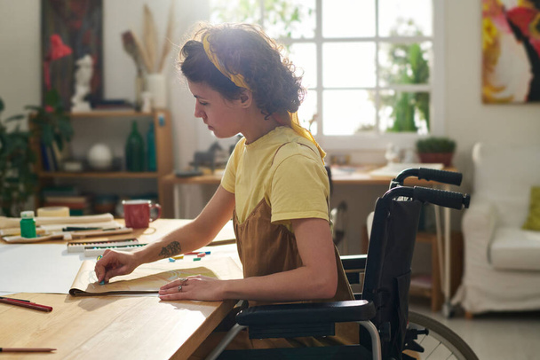 Молодая женщина с инвалидностью в инвалидной коляске создает новые рисунки с карандашами, сидя на рабочем месте в уютной домашней студии - Фото, изображение