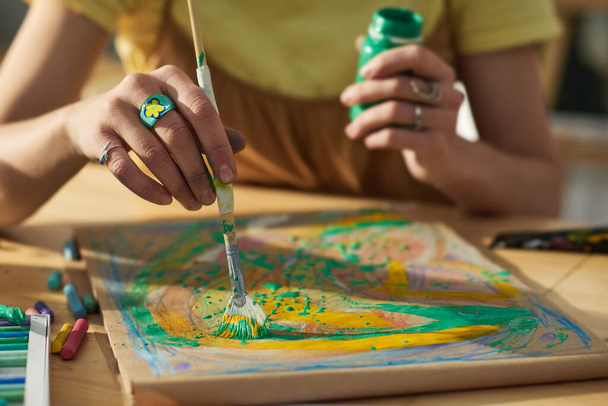Egy fiatal, festőecsettel és gouache-szal rendelkező kreatív nő kezéről készült kép, amint papíron új műalkotásokat készít, miközben a stúdióban ül a munkahelyén. - Fotó, kép