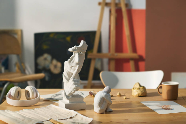 Arbeitsplatz moderner Kunsthandwerker mit Skulpturen aus Ton und Gips, Skizze auf Papier, Tasse und Kopfhörer gegen Malerei und Staffelei - Foto, Bild