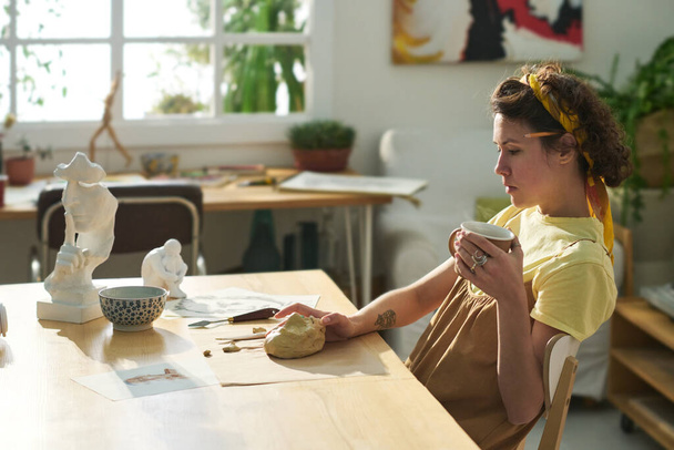 Молодая задумчивая ремесленница с чашкой чая думает о новых творческих идеях, сидя за столом с ручной работы фигурками и кусочком глины - Фото, изображение