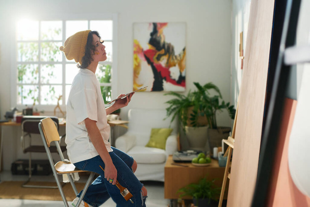 Νεαρή σκεπτική crfaftswoman με χρωματική παλέτα που δημιουργεί νέο αριστούργημα ενώ κάθεται στο στούντιο μπροστά σε ημιτελή έργα τέχνης - Φωτογραφία, εικόνα