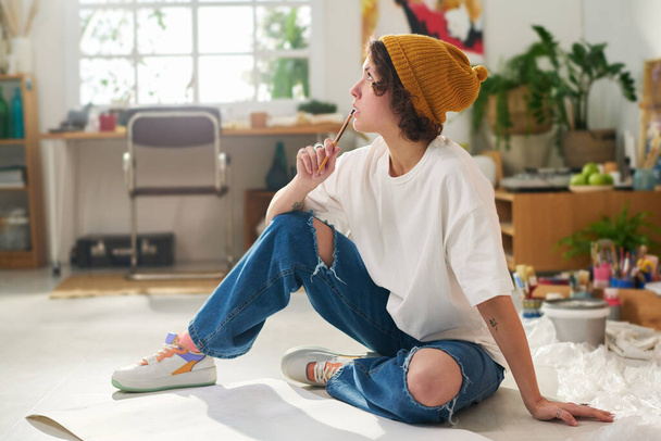 Νεαρή σκεπτική γυναίκα με casualwear κρατώντας μολύβι από το στόμα της, ενώ κάθεται στο πάτωμα με χαρτιά και σκέφτεται νέα έργα τέχνης - Φωτογραφία, εικόνα