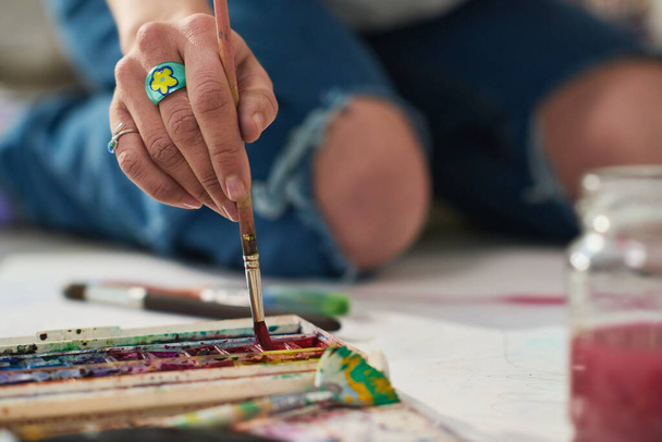 Focus op de hand van jonge creatieve vrouw met bijou ring zetten verfborstel in aquarel terwijl zitten op de vloer van de werkplaats en het creëren van kunstwerk - Foto, afbeelding