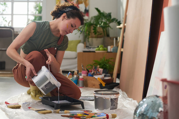 Молодая креативная художница заливает жидкую краску в пластиковый поднос перед тем, как начать работу над созданием нового шедевра в домашней студии - Фото, изображение