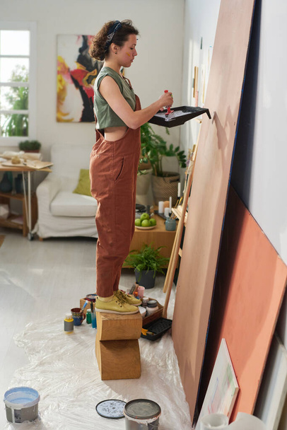 Боковой вид молодой творческой художницы в повседневной одежде, создающей новый шедевр, стоя перед панелью в домашней студии - Фото, изображение