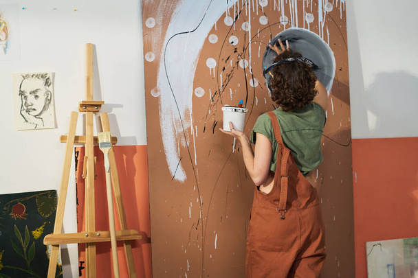 Achteraanzicht van jonge creatieve vrouw met verf die nieuw abstract kunstwerk creëert terwijl ze in haar atelier voor het paneel staat - Foto, afbeelding