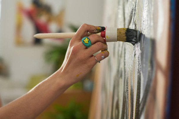 Рука молодой художницы с кистью во время создания новых работ в домашней студии или мастерской и часть ее шедевра - Фото, изображение