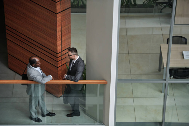 Teil des Interieurs eines modernen Geschäftszentrums mit zwei interkulturellen männlichen Angestellten in Formalbekleidung, die bei einem Treffen am Geländer diskutieren - Foto, Bild