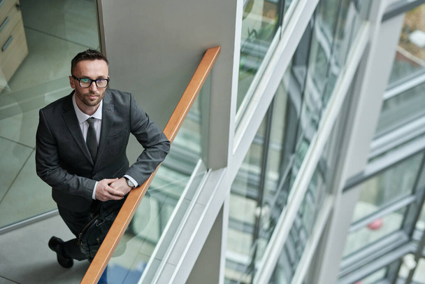 Νέοι αυτοπεποίθηση διευθύνων σύμβουλος σε formalwear στέκεται από διαφανή κιγκλίδωμα στο κέντρο του γραφείου και κοιτάζοντας κάμερα - Φωτογραφία, εικόνα