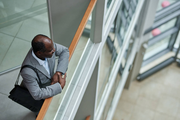 Above shot of mature African American broker with black handbag on shoulder standing by railing inside modern office center - Photo, image
