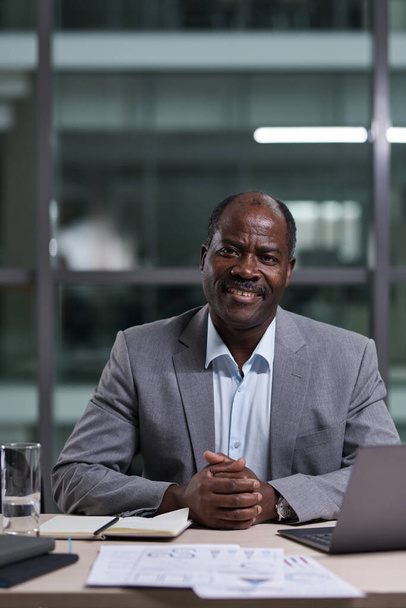 Успешный взрослый афроамериканец-директор бизнес-компании смотрит в камеру, сидя за компьютером на рабочем месте - Фото, изображение