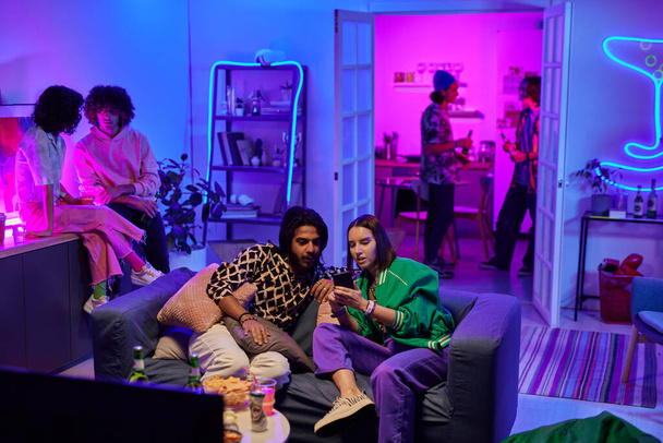 Молодая межкультурная пара просматривает новые фотографии в мобильном телефоне, сидя на диване на домашней вечеринке против своих друзей - Фото, изображение