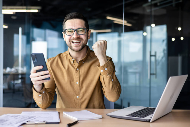 Portrait of a young happy businessman sitting in the office, holding a phone in his hand and looking at the camera. I received good news by message, a successful financial deal, a new project. - 写真・画像