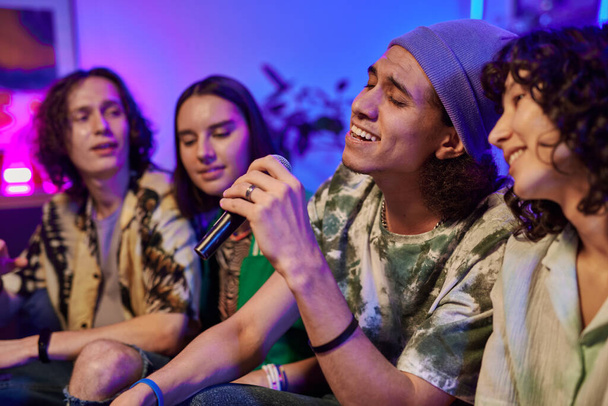 Selektywne skupienie się na młodym człowieku z mikrofonem śpiewającym karaoke ze swoją dziewczyną podczas zabawy w domu z przyjaciółmi w weekend - Zdjęcie, obraz