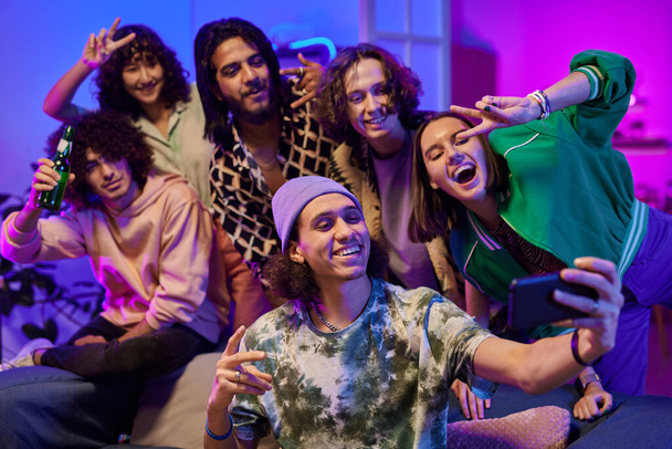 Grupo de jóvenes amigos interculturales alegres en ropa de casualwear tomar selfie en la fiesta en casa, mientras que de pie en la sala de estar iluminada por la luz de neón - Foto, Imagen