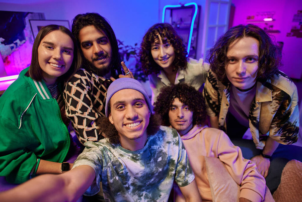 SIx młodych międzykulturowych mężczyzn i kobiet pozowanie do selfie przed kamerą podczas korzystania z home party w salonie oświetlony światłem neonowym - Zdjęcie, obraz