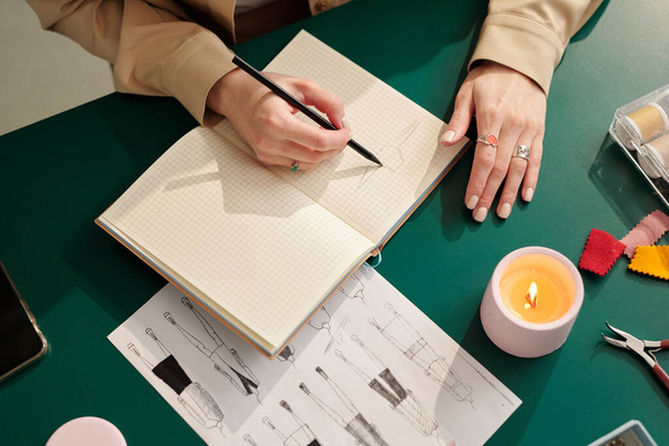 Πάνω γωνία της νέας δημιουργικής γυναίκας σχεδιάστρια μόδας με μολύβι πάνω από ανοιχτό σημειωματάριο σχέδια για νέα εποχιακή συλλογή - Φωτογραφία, εικόνα