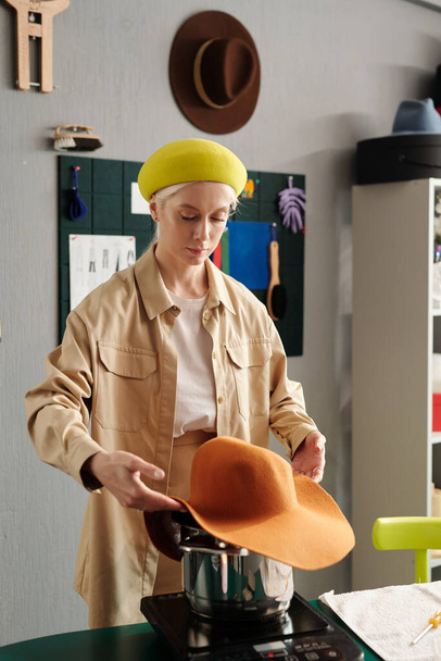 Jonge creatieve hoedenmaker in slim casualwear stand-by werkplek in design studio en stomend nieuw item over pan met kokend water - Foto, afbeelding