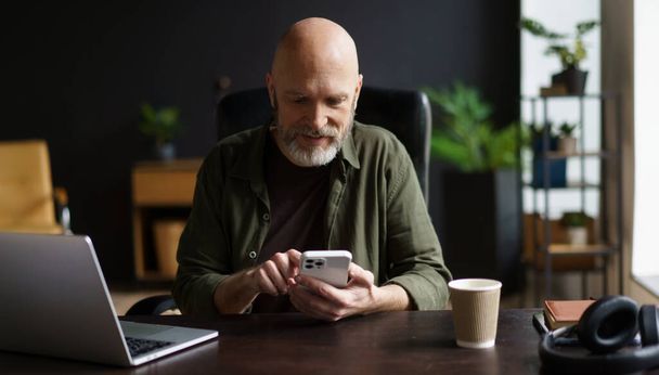 Happy and charismatic senior man who engrossed in text message on phone while taking break from work on laptop. Senior man, full of vitality, seen enjoying comfort of own house. . High quality photo - Photo, image