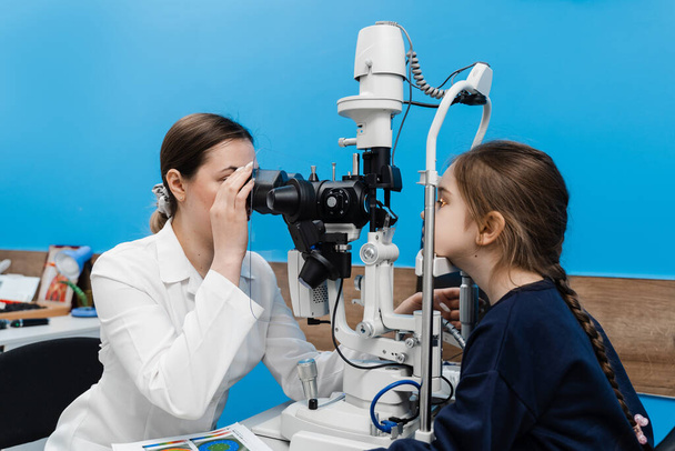 Детский офтальмолог с щелевой лампой исследует глаза и роговицу ребенка. Офтальмолог освещает глаз ребенка светом от щелевой лампы для диагностики глаз и роговицы - Фото, изображение