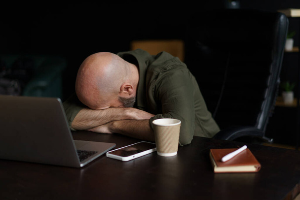 Tired mid-aged worker sleeping with head resting on table near laptop. Concept of age-related changes depicted, highlighting impact of exhaustion and fatigue on individuals as they navigate challenges - Foto, Imagen