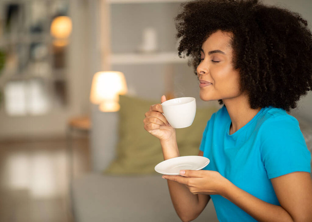 Positive millennial curly black lady hold cup of tea, enjoy aroma, good tasty, free time on sofa in cozy living room interior, copy space. Morning coffee break in evening, rest, relax at home - Photo, Image