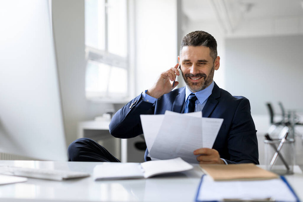 Middle aged businessman talking on cellphone and checking papers, sitting at workplace in office, man entrepreneur reading financial reports and making phone call to business partner, free space - Foto, afbeelding