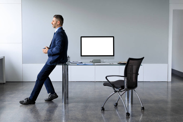 Hombre de negocios exitoso en ropa formal sentado apoyado en la mesa con maqueta de monitor de computadora en blanco, hombre mirando a la ventana, trabajando en la oficina, espacio de copia gratis, longitud de cuerpo completo - Foto, Imagen