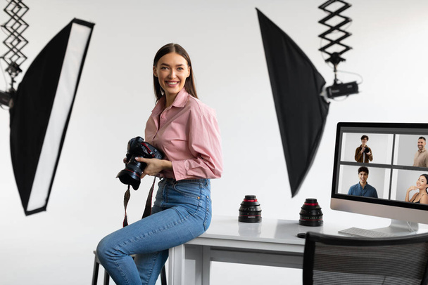 Щаслива творча фотографка леді сидить на робочому столі, тримаючи камеру DSLR, посміхаючись на камеру, працюючи з комп'ютером в сучасній фотостудії
 - Фото, зображення