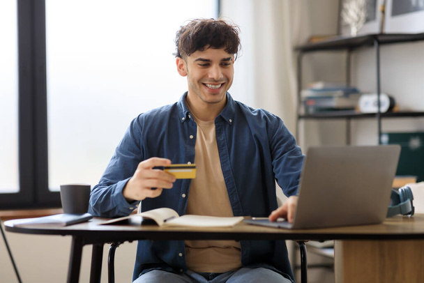 Online platební služba. Veselý arabský mladý muž nakupování pomocí notebooku a kreditní karty pro snadnou platbu sedí u stolu v kanceláři. Nabídka a koncepce elektronického obchodu. Selektivní zaměření - Fotografie, Obrázek