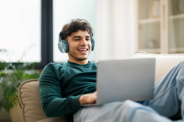 E-learning aanbod. Vrolijke Arabische Man typen en Websurfen op Laptop Draadloze oortjes dragen Zittend op de bank thuis. Studenten hebben plezier en leren Online met behulp van Computer - Foto, afbeelding