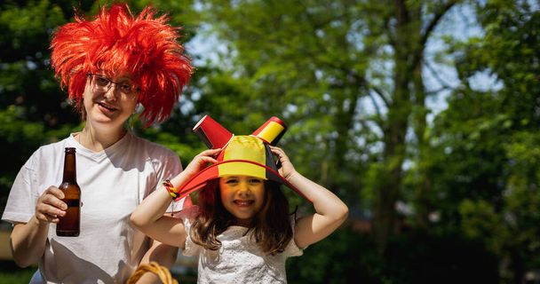Porträt der schönen und glücklichen kaukasischen Mutter in roter Perücke und ihrer kleinen Tochter mit belgischem Fahnenhut, die den belgischen Tag bei einem Picknick im Stadtpark an einem sonnigen Sommertag feiert, Nahaufnahme Seitenansicht - Foto, Bild