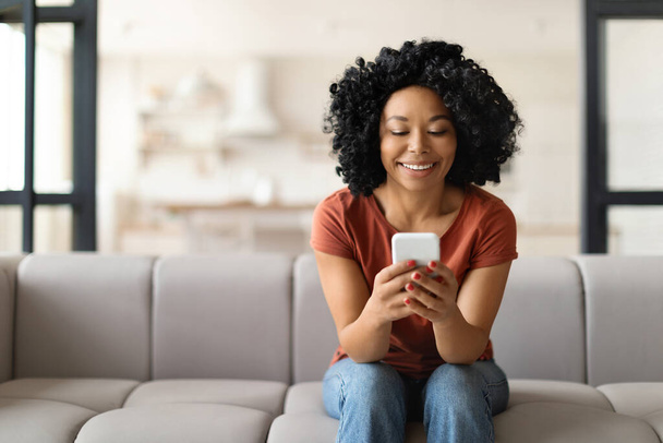Nuori hymyilevä musta nainen, jolla on älypuhelin, lepää kotona sohvalla, onnellinen afroamerikkalainen nainen istuu sohvalla ja käyttää matkapuhelinta, viestii ystävien kanssa tai selaa uutta sovellusta ostoksille - Valokuva, kuva