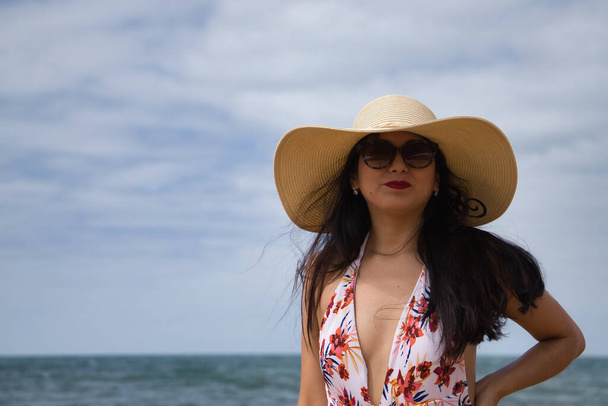 South American woman, young and beautiful, brunette with sunglasses, hat and swimsuit posing happy and smiling on the beach. Concept sea, sand, sun, beach, vacation, travel, summer. - Fotoğraf, Görsel