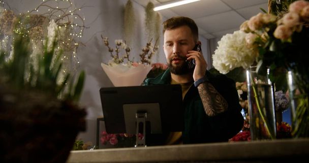 Férfi vállalkozó, tulajdonos, virágárus beszél telefonon, és veszi az ügyfél érdekében online digitális tabletta. Virágok a vázákban a virágüzletben. Virágzás, virágos kisvállalkozások és vállalkozói szellem. - Fotó, kép