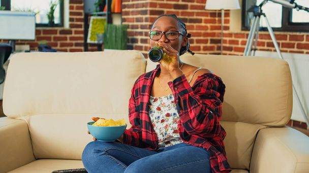 Smiling person holding bowl of snacks watching show, drinking alcohol and eating takeaway meal from multiple fast food orders. Young adult having burgers, fries, pizza and asian food at home. - Foto, imagen