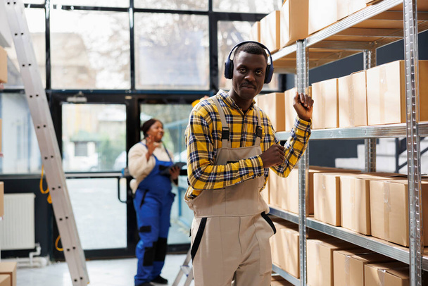 African american logistics manager dancing in factory storehouse and looking at camera. Young man wearing workwear listening to music and making hands movements in warehouse - Foto, Bild