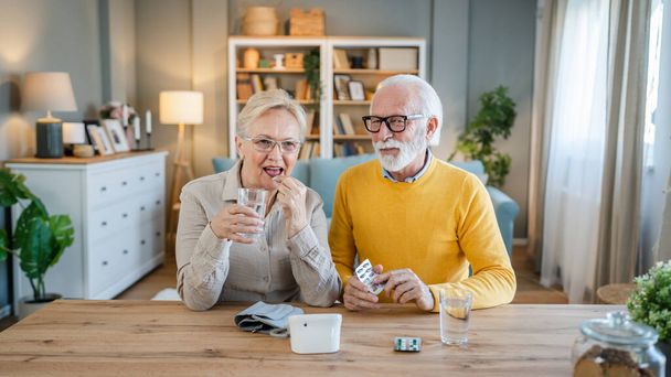 Senior couple woman take medicine or supplement vitamin treatment cure while her husband old caucasian man sit beside at the table at home real people family old people retirement and health concept - Photo, Image